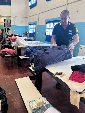 Un agente de policía con guantes blancos examina un bolso azul sobre una mesa en una habitación con paredes azules y blancas; otros agentes se ven al fondo