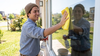 Hombre sonriente con gorro de lana y camisa azul limpia una ventana con un paño amarillo. Sostiene una botella rociadora con vinagre y agua.