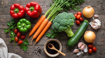 Vista superior de pimientos, zanahorias, brócoli, calabacín, cebollas, ajos, tomates y un recipiente con salsa de soya sobre madera.