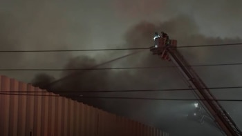 Imagen nocturna de bomberos en una escalera aérea rociando agua sobre un edificio envuelto en humo. Se ven líneas eléctricas aéreas horizontales cruzando el encuadre