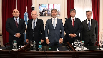 Cinco hombres de traje están de pie detrás de una mesa de madera en un recinto con cortinas rojas y una bandera argentina al fondo