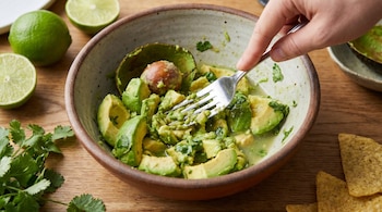 Una mano machaca aguacate en un cuenco rústico con un tenedor, junto a limas cortadas, cilantro y totopos sobre una superficie de madera clara.