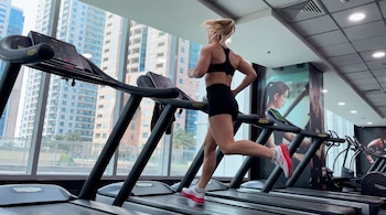 Mujer rubia corre en una cinta de correr dentro de un gimnasio. Detrás, edificios altos y palmeras se ven a través de ventanales. Otros aparatos de ejercicio.