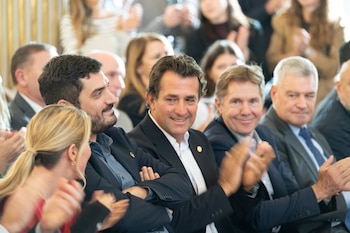 Grupo de hombres y una mujer de cabello rubio, vestidos formalmente, sentados y aplaudiendo. Varios sonríen. Fondo borroso con más asistentes