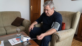 Hombre de mediana edad con cabello gris sentado en un sillón beige, midiéndose la presión arterial con un monitor. Un folleto de hipertensión y un vaso de agua están sobre la mesa.