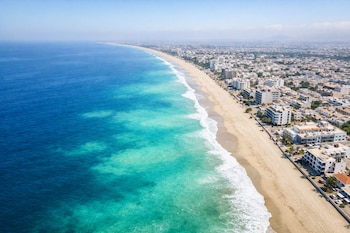 Vista aérea de una larga playa de arena dorada bordeada por un mar azul intenso con áreas turquesa. A la derecha, una ciudad costera con edificios bajos.