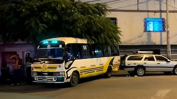 Una cúster blanca con amarillo estacionada de noche junto a un auto y bajo un árbol en una calle