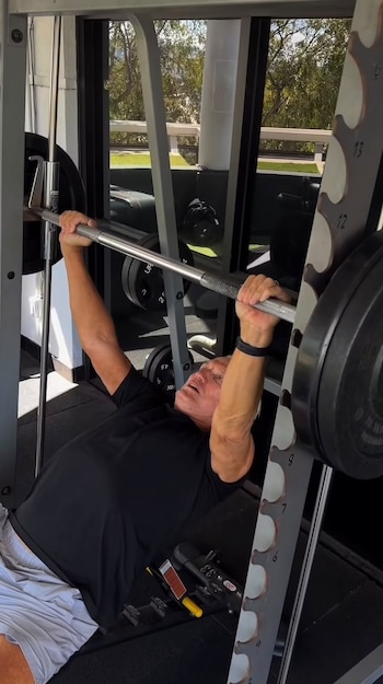 Dolph Lundgren acostado en un banco de gimnasio mientras sostiene una barra de pesas en una máquina guiada
