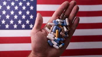 Primer plano de una mano sosteniendo un puñado de píldoras y cápsulas de diferentes colores y tamaños, con la bandera de Estados Unidos difuminada al fondo