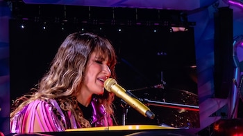 Primer plano de Hannah Pérez de Ha*Ash cantando en un micrófono dorado. Tiene cabello ondulado castaño y flequillo, viste una chaqueta rosa brillante con flecos