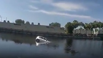 Un coche blanco se hunde en un canal de agua oscura; su parte trasera y techo están visibles. Edificios y árboles se distinguen al fondo