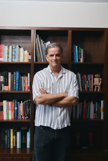 Un hombre con camisa a rayas blancas y negras posa con los brazos cruzados delante de una biblioteca de madera con estantes llenos de libros