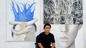Un hombre de piel morena con camiseta negra sentado, flanqueado por dos cuadros con figuras abrazadas y un árbol azul, y otro con árboles y parte de un rostro