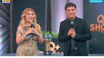 Gisela Valcárcel sostiene un micrófono y una carpeta junto a un hombre de traje negro frente a un trofeo en el set del programa Q+ Show