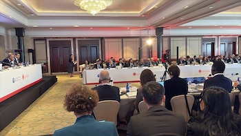 Personas sentadas en mesas de una sala de conferencias con micrófonos, auriculares de traducción y paneles con logotipos institucionales
