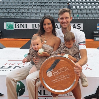 Un hombre sonriente, una mujer y dos bebés posan en una cancha de tenis con un trofeo de finalista de Internazionali BNL d'Italia. El fondo muestra asientos de estadio vacíos