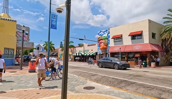 Una calle con edificios comerciales, un teatro, personas caminando, automóviles y una motocicleta en la vía, palmeras, postes de luz y un cielo azul con nubes