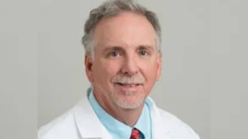 Primer plano de un hombre senior sonriente con cabello canoso, bigote y perilla, vistiendo una bata blanca, camisa azul y corbata roja