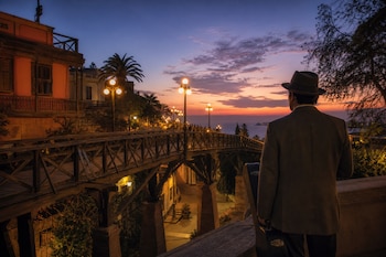 Un hombre de espaldas con sombrero sostiene un estuche de guitarra frente a un puente de madera iluminado por faroles al atardecer.