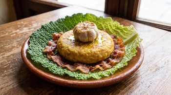Un plato de barro sobre una mesa de madera contiene una tortilla de patatas sobre una hoja de col, lonchas de panceta y un ajo encima.