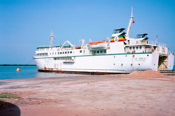 Un gran barco blanco llamado Le Joola con franjas verdes, amarillas y rojas atracado junto a un muelle de concreto bajo un cielo despejado