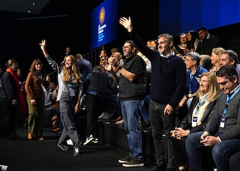 Grupo de personas sonriendo y aplaudiendo en un evento, con algunos brazos levantados y un cartel azul con el logo del sol en el fondo