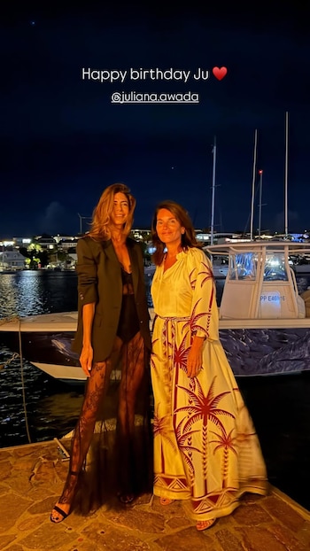 Dos mujeres, una con blazer negro y la otra con vestido blanco estampado, posan sonrientes en un muelle por la noche con barcos y luces al fondo