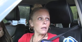 Una mujer rubia con el pelo recogido y grandes pendientes de aro, vestida de rojo, mira hacia un lado con expresión de sorpresa o angustia. Un micrófono de noticias está en primer plano