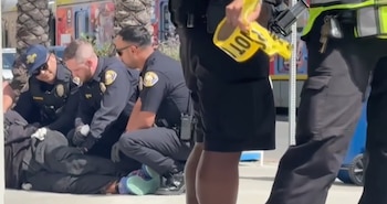 Varias personas vestidas de policía deteniendo a un hombre tendido en el suelo en un área al aire libre con palmeras y un camión de comida de fondo