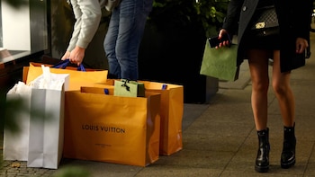 Una persona con bolsas de compra Louis Vuitton y Gucci, y otra cargando bolsas, caminando en una acera. Bolsas naranjas de Louis Vuitton y una verde de Gucci en primer plano