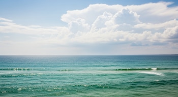 Océano de tonos azul verdosos con pequeñas olas en primer plano, horizonte visible, y cielo azul claro con grandes nubes blancas y grises.