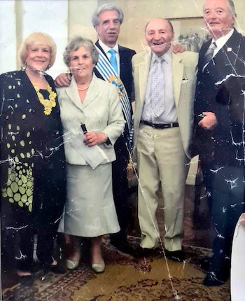 Cinco personas, tres hombres y dos mujeres, posan sonriendo. Un hombre usa una banda celeste y blanca. Fondo claro con alfombra estampada. La imagen presenta desgaste