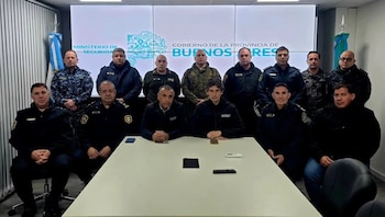 Catorce hombres con uniformes de seguridad y ropa formal posan junto a una mesa frente a una pantalla institucional en una sala