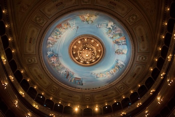 Vista cenital de la cúpula del Teatro Colón: mural circular con figuras, instrumentos y paisajes, y una gran araña dorada central. Balcones iluminados