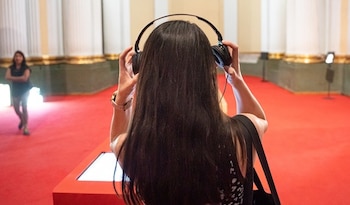 Vista trasera de una persona con cabello largo oscuro, ajustándose auriculares negros frente a una pantalla brillante, sobre una alfombra roja en una galería