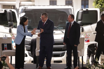 Varios funcionarios caminan junto a una hilera de camiones blancos con emblemas oficiales de Guatemala y Taiwán en un estacionamiento