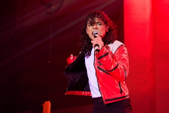 Gabriela Villanueva, con cabello rizado y chaqueta roja brillante, canta con un micrófono en mano, iluminada por luces rojas en el escenario
