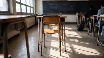 Imagen realista de una banca de madera y metal vacía en una clase de preparatoria, con luz solar incidiendo desde grandes ventanas a la izquierda. Un pizarrón negro y otros pupitres ocupados se ven al fondo.