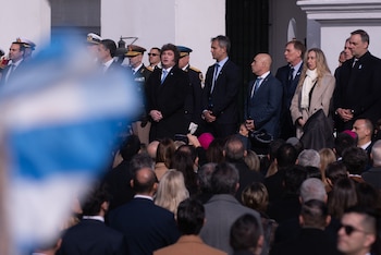 Presidente y funcionarios en trajes oscuros y uniformes militares, junto a civiles, de pie en un evento. Parte de una bandera argentina en primer plano