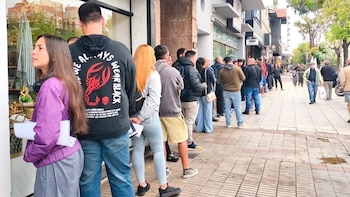 Larga fila de personas de pie en una acera de la ciudad, esperando frente a una tienda con ventanas, con edificios y árboles al fondo
