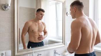 Hombre sin camiseta frente al espejo de un baño con la mano colocada sobre el abdomen.