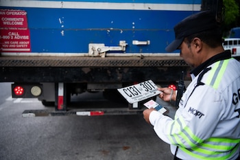 Un agente con chaleco reflectante sostiene una placa de matrícula provisional y unos documentos junto a la parte trasera de un camión azul