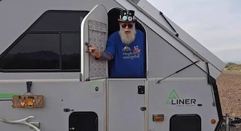 Hombre de barba blanca con sombrero y gafas oscuras en la puerta de una caravana gris de marca Aliner