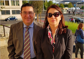 Un hombre con traje y corbata junto a una mujer con gafas de sol y pañuelo. Fondo urbano con vehículos, edificios y vegetación