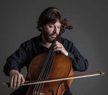 Hombre de camisa negra y barba que sostiene un violonchelo y apoya el arco sobre las cuerdas ante un fondo gris
