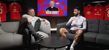 Wayne Rooney y Bruno Fernandes sentados en sofás, con una pantalla detrás mostrando "The Wayne Rooney Show" y camisetas rojas del Manchester United colgadas