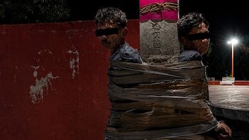 Dos hombres con ojos censurados, uno con 'RATA' en la frente, están atados con cinta adhesiva a un poste de concreto, al aire libre y de noche