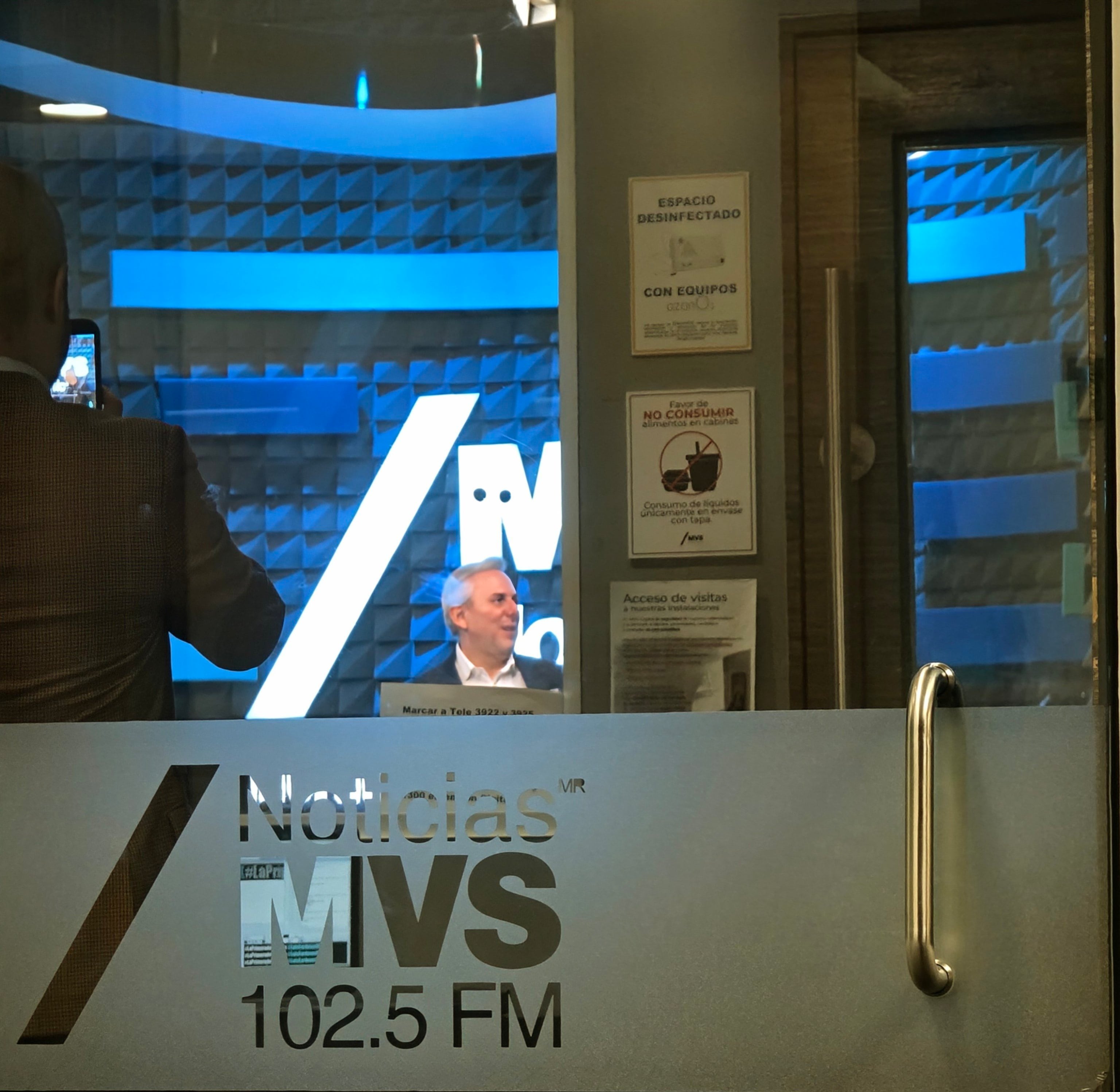 Luis Cárdenas sonríe durante la emisión de su último programa radiofónico, visto a través de la puerta de un estudio de Noticias MVS 102.5 FM. (X: Mejía Salvador)