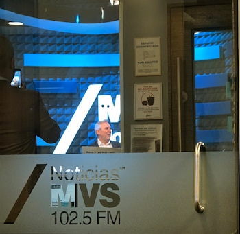 Hombre dentro de estudio de radio azul, visible a través de puerta de vidrio con logo Noticias MVS 102.5 FM. Otra persona toma una foto con un celular