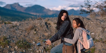 Cazzu, una mujer de cabello oscuro largo, en bicicleta con una niña detrás. Están en un paisaje rocoso y árido con montañas distantes